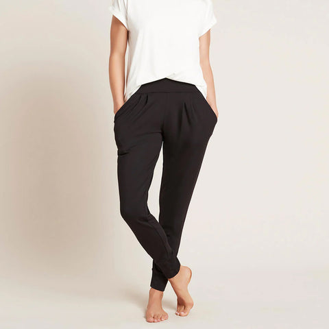 A model wearing black, lightweight lounge pants made of organic bamboo, featuring a draped fit with a wide waistband, tapered leg and two side pockets, showing the front with a hand in pocket.