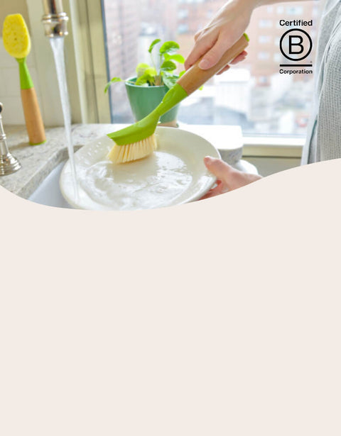 Person washing a plate with a green dish brush in a kitchen.
