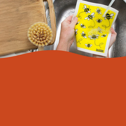 Hand holding a yellow sponge with bee pattern next to a wooden cutting board and sink.