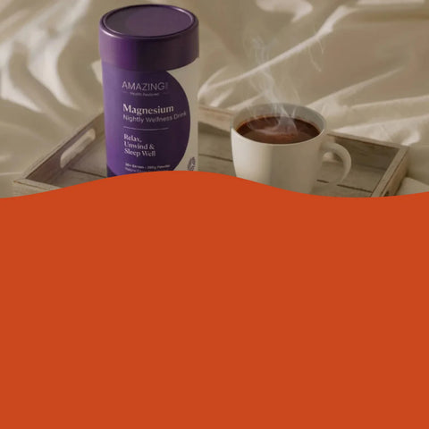A purple cylindrical canister of Amazing Magnesium supplement placed on a wooden tray beside a white mug of hot tea with visible steam, set against draped cream fabric. A large solid orange shape overlays the lower portion of the frame.