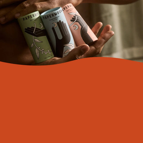 Three cylindrical paperboard packaging tubes held in hands against a warm neutral background, with a large solid orange shape obscuring the lower half of the frame. The tubes are labeled PAPERMOUS.