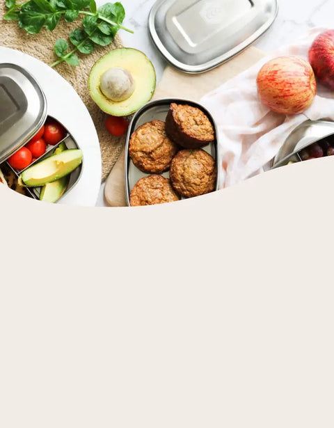 A styled flat-lay of stainless steel bento lunch boxes with food items including avocado, tomatoes, greens, and muffins, arranged on a light surface with a large blank beige space below.
