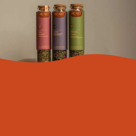Three glass canisters of Heart brand herbal tea are shown standing upright against a neutral background, with a large solid orange graphic overlay covering the bottom half of the frame.