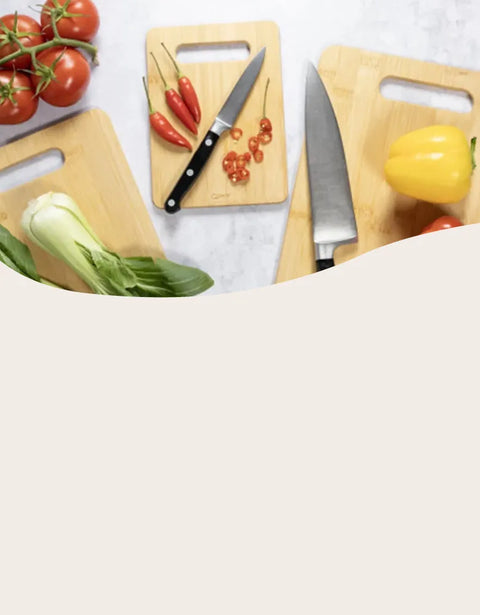Three wooden cutting boards with integrated handles are arranged on a light marble surface, styled with kitchen knives, tomatoes, red chilies, sliced red peppers, a yellow bell pepper, and a green cabbage. A large beige graphic overlay covers the bottom half of the frame.