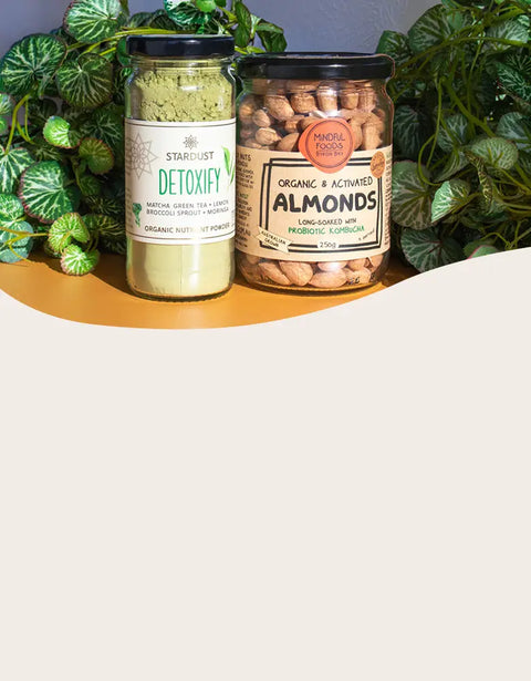 Two jars of health foods: a glass jar of Starlust Detoxify powder on the left and a glass jar of Organic & Activated Almonds on the right, displayed on a wavy tan surface with green foliage in the background.