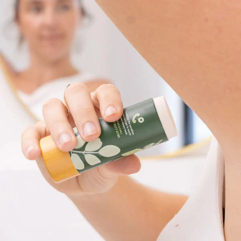 A close-up of a hand holding a green and yellow stick deodorant in front of a blurred mirror showing a woman's face.