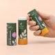 Two tubes of natural stick deodorant displayed on a warm peach background. One tube is held by a hand, one is standing upright.