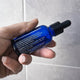 Hand holding a blue dropper bottle with text against a gray tiled wall.