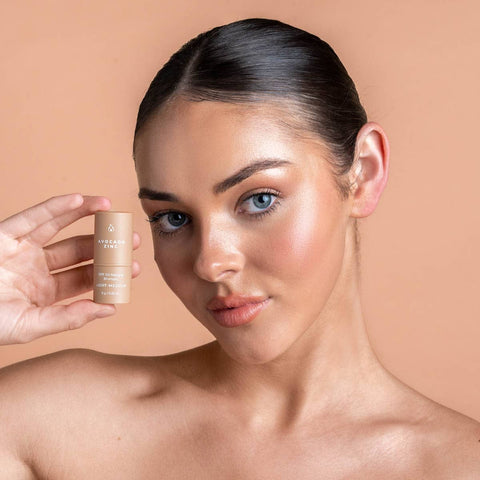 Model holding Avocado Zinc SPF Natural Bronzer next to her face, which has been perfectly made up to look natural, glowy and bronzed. 