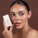 Woman holding glow drops next to her perfectly made-up face with a brown background