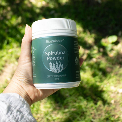 Hand holding a container of BioBalance Spirulina Powder against a natural background.