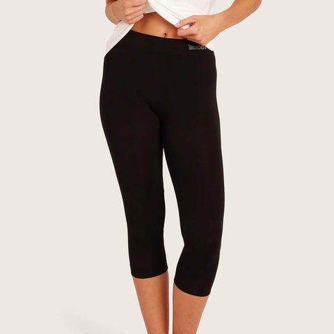 Black capri leggings worn by a model, shown from the waist down against a light beige studio background. The model is wearing a white top and pulling the waistband slightly outward with both hands.