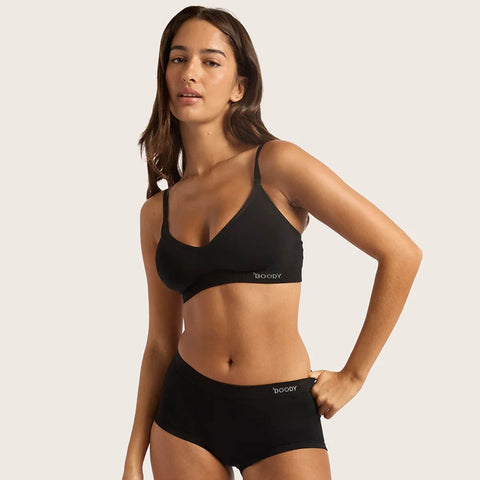 A woman modeling a black two-piece lingerie set against a solid light beige background. The set includes a bralette with thin straps and matching high-waisted briefs, both featuring the brand name 'BOODY' printed in white on the front.