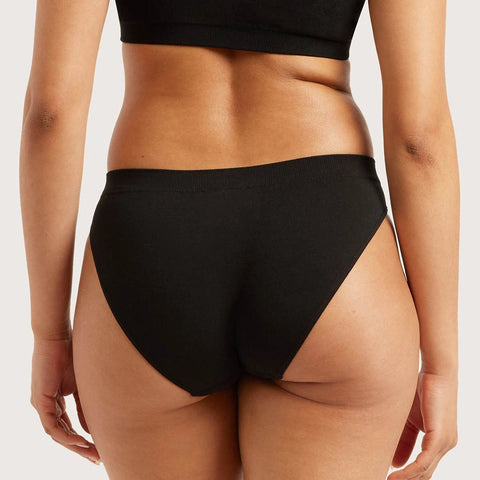 Rear view of a model wearing black high-waisted briefs with a side cutout, paired with a black sports bra against a light gray studio background.