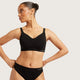 A woman modeling a black bralette and matching black underwear bottoms against a light gray studio background. The brand name 'boody' is visible on the waistband of the underwear.