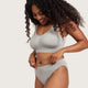 A woman modeling a light gray two-piece lingerie set consisting of a scoop-neck bralette and matching high-waisted briefs, against a solid off-white background.