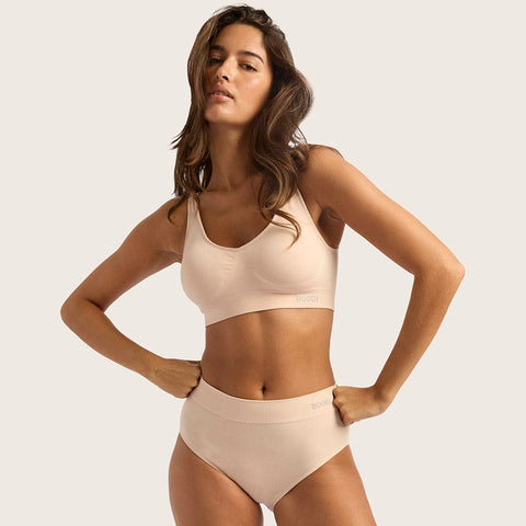 A woman modeling a matching beige/nude two-piece underwear set consisting of a wireless bra and high-waisted briefs, against a solid light beige background.