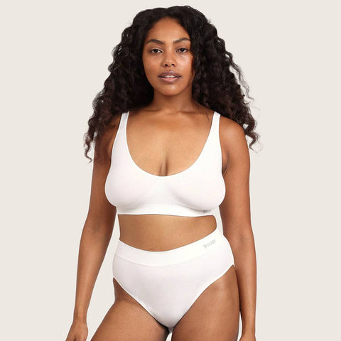 A woman modeling a white two-piece bra and panty set against a solid light beige background.