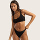 A woman modeling a black two-piece bikini set against a light beige studio background. The set includes a scoop-neck bikini top and matching high-cut bikini bottoms, both with small white 'BOODY' branding on the side.