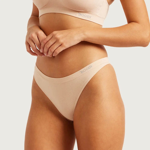 A close-up of a model wearing a light beige/peach high-waisted brief and matching bra from Booby, with the brand name visible on the waistbands.
