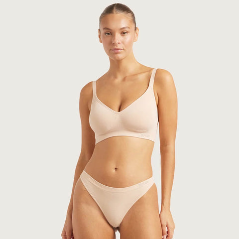 A model wearing a light beige two-piece lingerie set consisting of a bralette and matching high-waisted briefs, photographed against a solid light gray background.