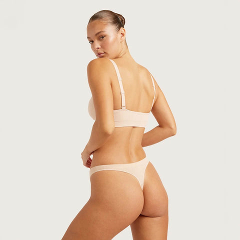 A model wearing a light beige two-piece lingerie set, featuring a bralette with adjustable shoulder straps and matching high-cut briefs, shown from a three-quarters back view against a solid light gray background.