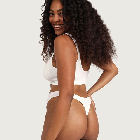 A woman with long, curly dark hair is shown from the back wearing a white two-piece bikini set against a plain light gray studio background.