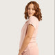 A woman with wavy brown hair wearing a light pink scrub top and matching scrub pants, shown from a side-back angle against a solid light gray background.