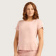 A woman with wavy brown hair wearing a light pink short-sleeve crewneck top and matching pink bottoms, photographed against a plain light gray studio background.