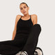 A woman wearing a black tank top and black pants, sitting against a light gray studio background.
