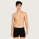 A male model wearing black boxer briefs, standing against a light gray studio background.