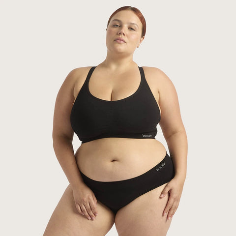 A plus-size model wearing a black two-piece lingerie set consisting of a scoop-neck bralette and matching high-waisted briefs, shown against a solid light gray background.
