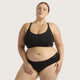 A plus-size model wearing a black two-piece lingerie set consisting of a scoop-neck bralette and matching high-waisted briefs, shown against a solid light gray background.