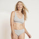 A smiling blonde woman wearing a light gray two-piece underwear set, consisting of a scoop-neck bralette and matching high-waisted briefs, against a plain off-white studio background.