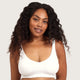A woman with long, curly dark hair wearing a white wireless bralette, photographed against a solid light gray background.