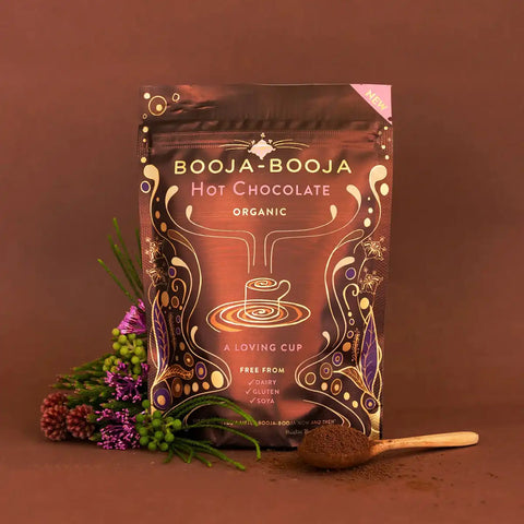 A stand-up pouch of Booja-Booja Hot Chocolate Organic coffee is displayed on a warm brown background, styled with a small wooden spoon of dark chocolate powder and dried flowers on the left.