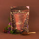 A stand-up pouch of Booja-Booja Hot Chocolate Organic coffee is displayed on a warm brown background, styled with a small wooden spoon of dark chocolate powder and dried flowers on the left.