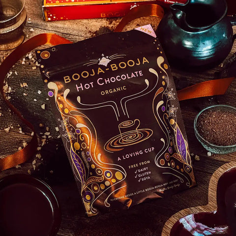 A bag of Booja-Booja Organic Hot Chocolate is displayed on a rustic wooden table surrounded by decorative elements like a dark teal teapot, a heart-shaped dish, and scattered chocolate powder.