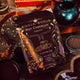 A bag of Booja-Booja Organic Hot Chocolate is displayed on a rustic wooden table surrounded by decorative elements like a dark teal teapot, a heart-shaped dish, and scattered chocolate powder.
