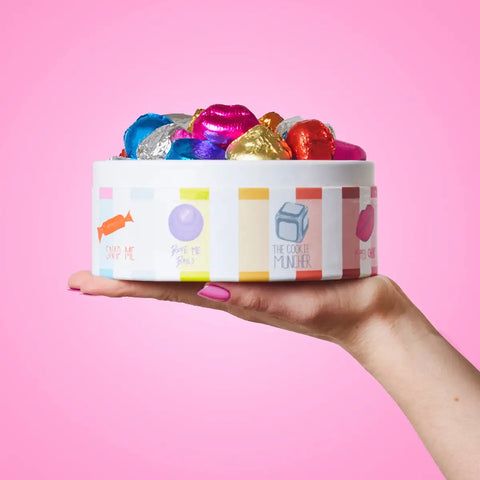 A hand with pink nail polish holds a white cylindrical box filled with colorful foil-wrapped chocolates against a solid pink background. The box features whimsical illustrations and text.