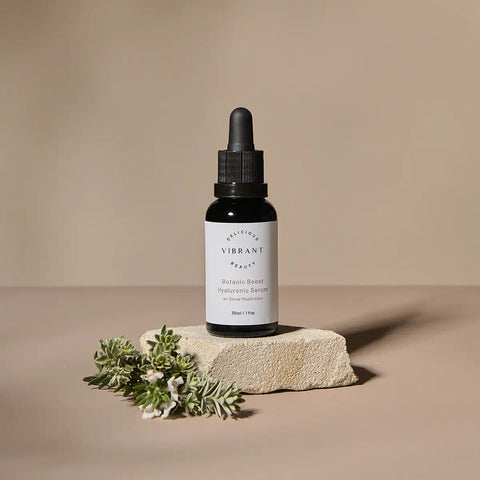Small glass dropper bottle labeled 'Botanic Boost Hyaluronic Serum' stands on a textured stone slab with green herb sprigs in the foreground against a warm beige studio background.