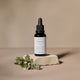 Small glass dropper bottle labeled 'Botanic Boost Hyaluronic Serum' stands on a textured stone slab with green herb sprigs in the foreground against a warm beige studio background.