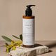 A bottle of Vibrant Beauty Gentle Cleansing Cream stands on a textured stone slab with yellow flowers and green leaves against a warm beige background.