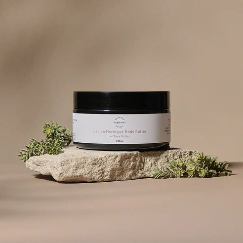 A jar of Lemon Meringue Body Butter sits on a textured stone slab with small green leaves, set against a warm beige background.