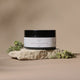 A jar of Lemon Meringue Body Butter sits on a textured stone slab with small green leaves, set against a warm beige background.