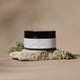 A 250ml jar of Luxe Rose Body Butter sits on a textured stone slab, flanked by small green leaves, against a warm beige background.