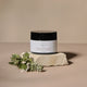 Night Radiance Sleep Mask jar in a black and white container, styled on a textured stone block with green leaves against a warm beige background.