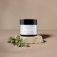 A jar of Night Rejuvenation cream sits on a textured stone slab with small green leaves in the foreground, set against a solid beige background.