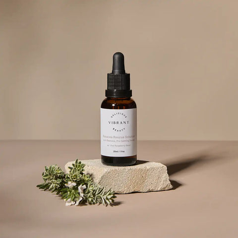 A bottle of Rosacea Rescue Solution sits on a textured stone slab with green rosemary sprigs, set against a warm beige background.