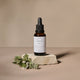 A bottle of Rosacea Rescue Solution sits on a textured stone slab with green rosemary sprigs, set against a warm beige background.